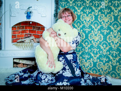 Happy young woman sitting near fireplace at home and hugging a white Teddy bear Stock Photo