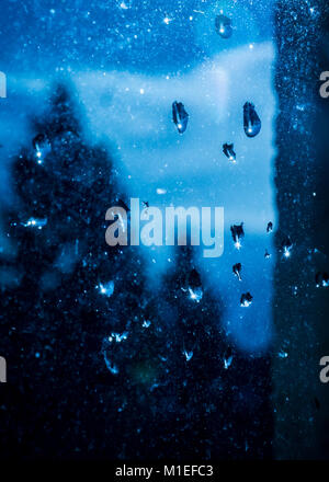Raindrops on a (rather dirty) window. Blue dramatic sky in the background. With reflection of trees in the droplet. Stock Photo