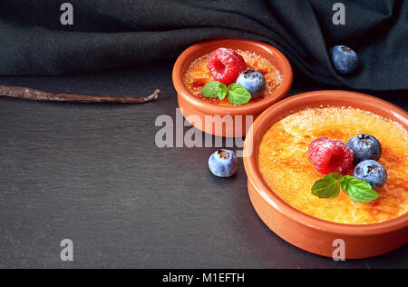 Food background with text space. Creme brulee (cream brulee, burnt cream) with raspberry and blueberry powdered with sugar on dark stone background. Stock Photo