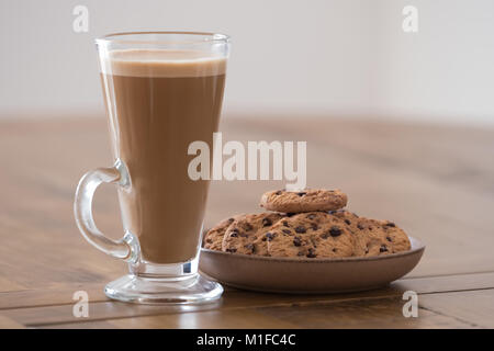 A tall glass of caffe latte and some chocolate chip cookies on a round plate all placed on a oak wooden table. Stock Photo