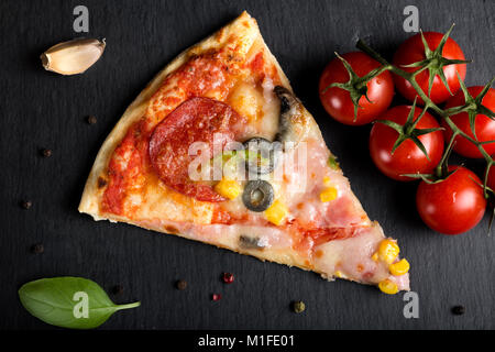 Slice of Italian Capriciosa pizza on dark slate with tomatoes and herbs Stock Photo