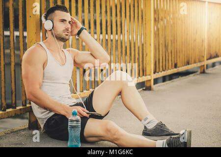 Fitness training. Young sportsman take a break after training. He siting on the bridge and listening to music with headphones. Fitness, sport, lifesty Stock Photo