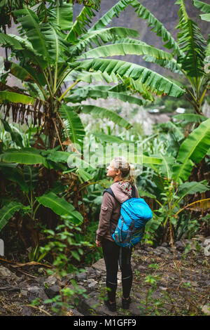 Blond yong women with blue backpack admire banana plantation on the trekking route to paul valley on santo antao island, cape verde Stock Photo