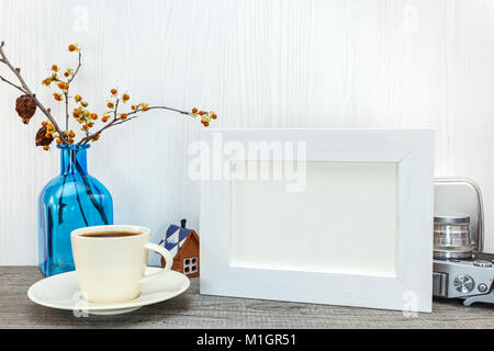 empty white photo frame, cup of coffee, blue glass vase with dried flowers on wooden background Stock Photo