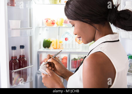 Close-up Of Young African Woman Writing On Spiral Book Near Open Refrigerator Stock Photo