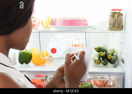 Close-up Of Young African Woman Writing On Spiral Book Near Open Refrigerator Stock Photo