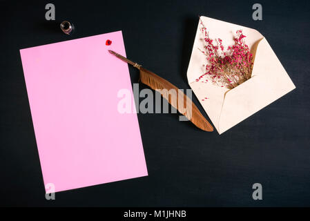 Open envelope full with red flowers, an antique feather pen on a pink paper note with a red ink splash on a black wooden background. Stock Photo