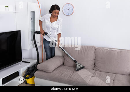 Happy Young African Woman Cleaning Sofa With Vacuum Cleaner At Home Stock Photo