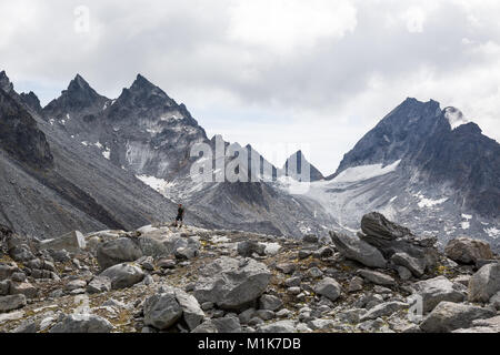 A hiker with his hands in his pockets stands on a rocky hill below an alpine scene of rocky peaks, glacier ice and scree slopes Stock Photo