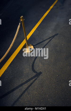 A gold rope barrier and stand on a road with a single yellow line and shadow - everyday special vip odd quirky minimal Stock Photo