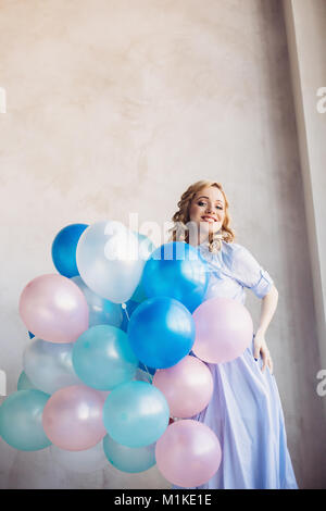 Blonde woman in light blue dress with blue and pink baloons stay in front of light wall Stock Photo