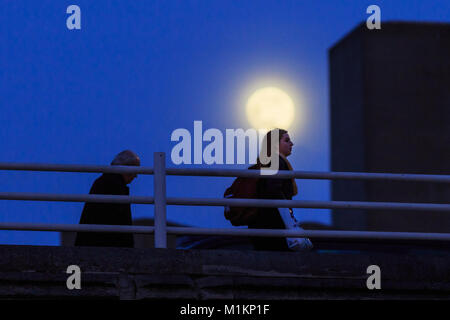 South Bank, London, UK. 30th Jan, 2018. A Waxing Gibbons Moon is visible in clear skies, as people walk across Waterloo Bridge and along the silhouetted National Theatre, on the night before the blue super moon which will be visible in the UK. Credit: Imageplotter News and Sports/Alamy Live News Stock Photo