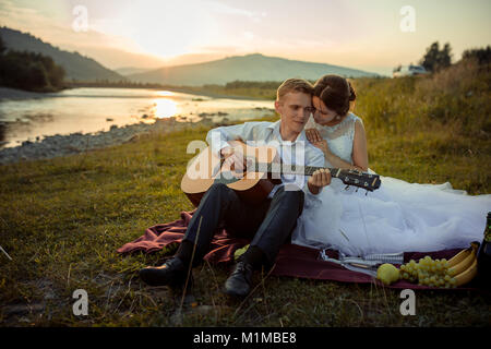 Attractive groom plays the guitar and his charming bride enjoys it during their picnic on the river bank during the sunset. Stock Photo