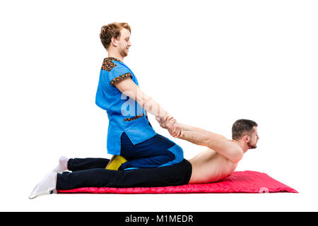 a young handsome man doctor masseur in a blue suit makes a traditional pose of takia massage to a guy lying on a living on a carpet rug. White isolate Stock Photo