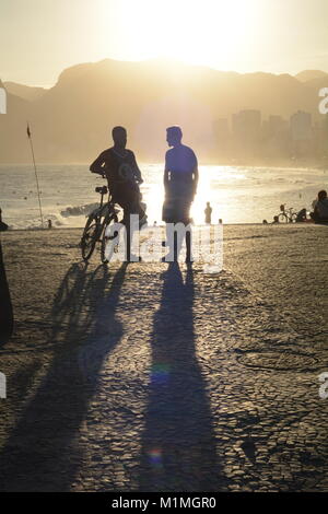Two friends chatting on Ipanema beach in Rio as the sun sets behind the mountains Stock Photo