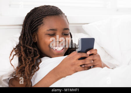 African Girl Lying On Bed Under The Blanket Using Cell Phone Stock Photo