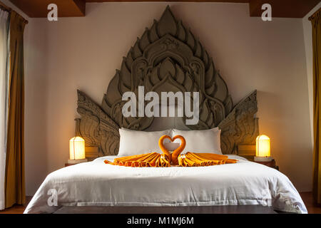 A full-size bed with a typical Thai style ornamented headboard and on the bed are two folded towel swans. Stock Photo