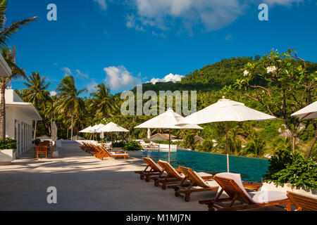 A perfect holiday photo of empty deck chairs and patio umbrellas in between, lined up in front of an infinity pool on a tropical island. Stock Photo