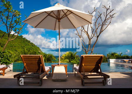 A typical holiday image of two deck chairs next to each other under the shade of a patio umbrella in front of an infinity pool in a tropical paradise. Stock Photo