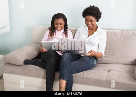 Smiling African Mother And Daughter Sitting On Couch Using Laptop And Digital Tablet Stock Photo