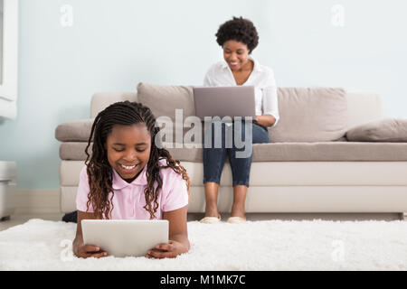 Girl Lying On Carpet Using Digital Tablet And Her Mother Using Laptop In Foreground Stock Photo