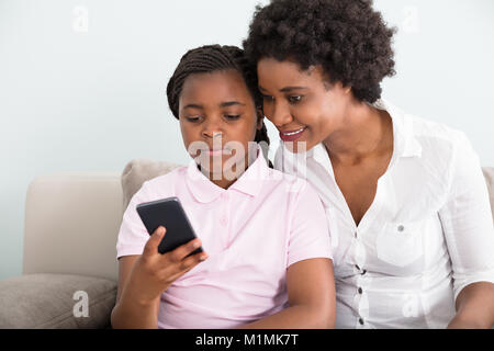 Mother And Daughter Sitting On Couch Looking At Smart Phone Stock Photo