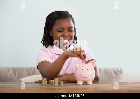 Portrait Of An African Girl Inserting Coins In Pink Piggy Bank On Table Stock Photo