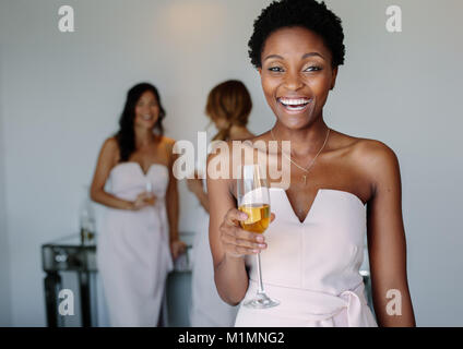 Beautiful young bridesmaid having wine on wedding day. Smiling african woman having drinks in a hotel room. Stock Photo