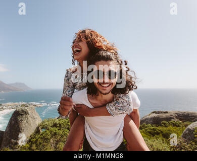 Smiling man giving piggyback ride to his girlfriend on the top of a mountain. Happy young couple enjoying life. Stock Photo