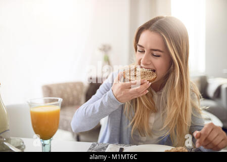 Pretty young teenage girl taking a bite of a healthy brown wholegrain roll as she enjoys a healthy breakfast at home with a glass of fresh orange juic