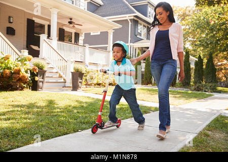 Mother Walks With Son As He Rides Scooter Along Sidewalk Stock Photo