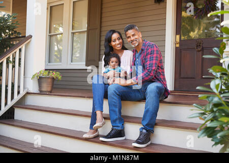 Family With Baby Son Sit On Steps Leading Up To Porch Of Home Stock Photo