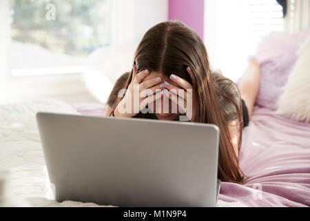 Upset teen girl lying on bed with head in hands using laptop Stock Photo
