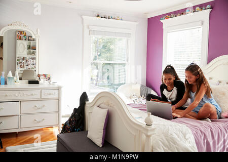 Two girlfriends in bedroom sitting on bed using a laptop Stock Photo