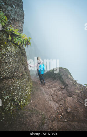 Hiker women with backpack walking down the steep slope of the rock in the foggy mountains. Stock Photo