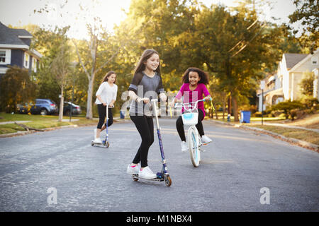 Three girls riding down the street on scooters and a bike Stock Photo