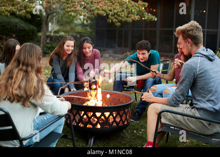 Teenage friends sit round a fire pit toasting marshmallows Stock Photo