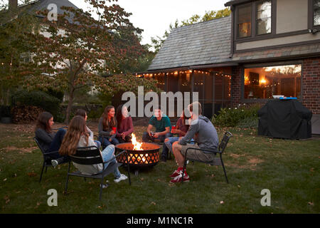 Teenagers sit talking around a fire pit in a garden at dusk Stock Photo