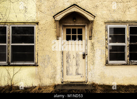 A close up of a cracked and faded exterior cement wall of a home with a door with wood awning above and a window on both sides Stock Photo