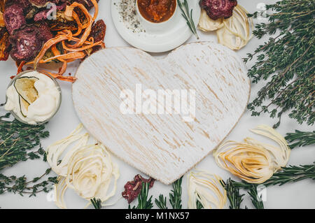 Homemade colorful chips of different fresh vegetables - beets, sweet potatoes, carrots, cucumber, onions, rosemary and thyme on a light background around the white Board in the shape of a heart. Stock Photo