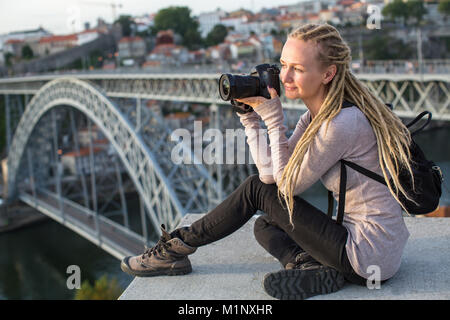 Young blond woman sitting with camera near Dom Luis I bridge across the Douro river in Porto, Portugal. Stock Photo