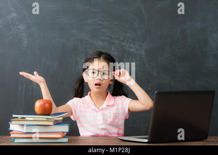 pretty attractive little girl children using computer studying having problem in chalkboard background and face to camera showing confused emotion. Stock Photo