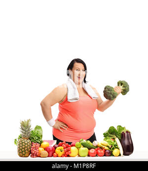 Overweight woman with a towel and a broccoli dumbbell behind a table with fruit and vegetables isolated on white background Stock Photo