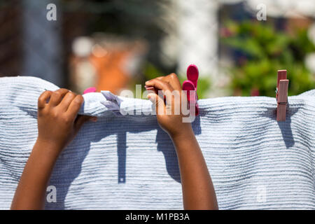 A young girl playing with clothes pegs and hanging a laundry blanket on a washing line in the garden. Stock Photo