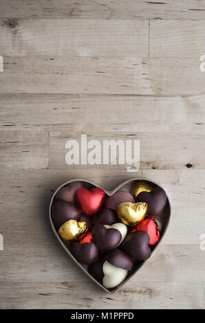 Overhead shot of open heart shaped box containing a variety of home made chocolates on old, worn light wood plank table. Stock Photo