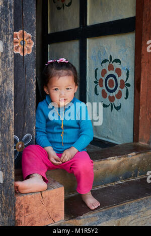 Young little girl sitting at doorway of house wearing blue top and pink bottoms in Bhutan Stock Photo
