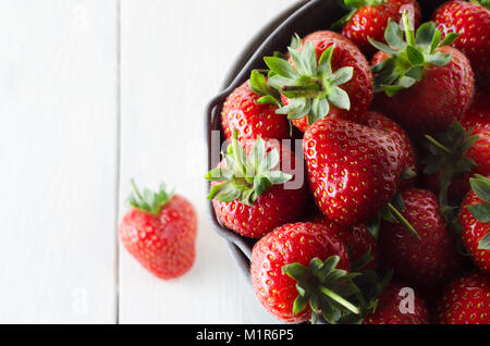 Overhead close up of a metal bucket, filled with red strawberries. One strawberry has fallen on white wood planked table below. Stock Photo