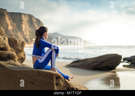 woman wearing a blue summer wetsuit sitting the on rocks watching the waves. Stock Photo