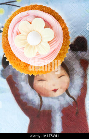 home made cupcake cake with strawberry pink frosting icing and rice paper edible daisy flower on top on serviette with girl holding hands up Stock Photo