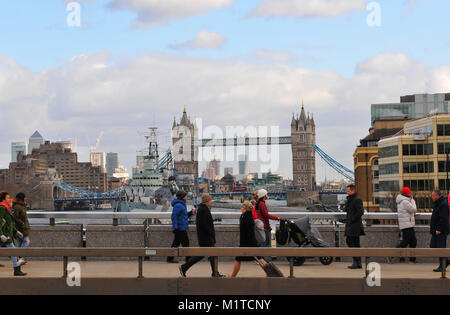 a number of people tourists and commuters walking over London bridge with tower bridge and HMS Belfast in the background over the river Thames London. Stock Photo